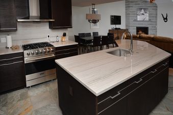 A modern kitchen with a large island, stainless steel appliances, dark wood cabinetry, and a view of a dining area and living room with a fireplace.