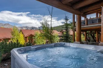 An outdoor hot tub with bubbling water, set against a backdrop of mountains and rustic log cabins with red roofs, surrounded by trees.
