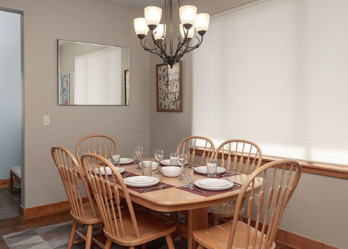 A bright dining area features a wooden table set with white dishes and clear glasses, surrounded by light wooden chairs, illuminated by an overhead chandelier.
