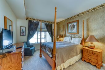 A bedroom with a four poster bed, a dresser with a television on top of it, a side table with a lamp, and a window with a view of a snowy mountain landscape.