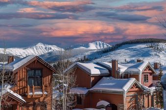 Snow-covered rustic wooden houses of Hygge Hideaway with majestic snowy mountains and a pink sunset in the background.