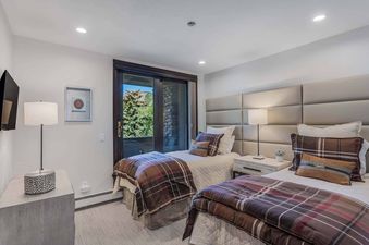 A serene twin bedroom at Woodrun Place 40 with two beds, plaid comforters, a shared nightstand with a lamp, a wall-mounted TV, and a sliding glass door leading to a balcony with mountain views.