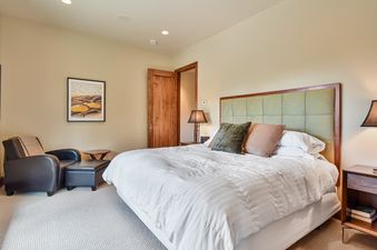 A cozy bedroom in Sky Villa features a king-sized bed with a green padded headboard, a dark leather armchair with an ottoman, and warm lighting, all set on light-colored carpeting.