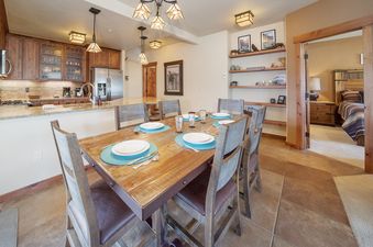 The dining area and kitchen of Cozen Pointe I201 are shown, featuring a wooden dining table set for six, surrounded by light wooden chairs with dark cushions. The kitchen has wood cabinetry and a large island with bar seating.