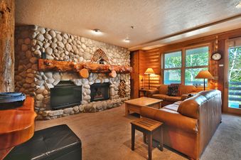 A cozy living room in Comstock 203 featuring a large stone fireplace, comfortable leather sofas, and views of green trees outside large windows.