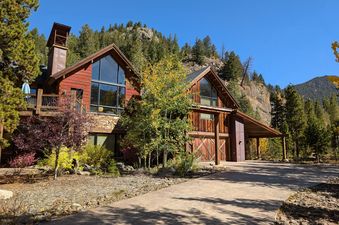 A large, multi-story home with red siding and stone accents is nestled into a forested mountain landscape under a clear blue sky.