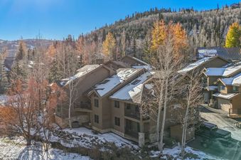 A drone shot of the Retreat on Deer Valley Dr, a multi-level building with snow on its roof, surrounded by trees with autumn foliage and some snow on the ground, against a clear blue sky and mountains in the background.