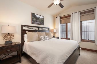 The bedroom features a comfortable king-sized bed with a wooden headboard, two bedside tables with lamps, and large windows with blinds and curtains.