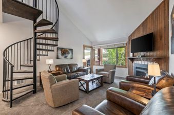 A spacious and inviting living room at Snow Flower 79 Park City, featuring comfortable leather seating, a spiral staircase, and a large window overlooking a verdant outdoor landscape.