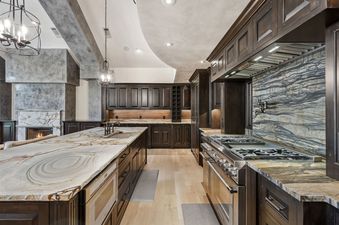 A luxury kitchen with dark wood cabinets, a large marble island, stainless steel appliances, and a blue-grey marble backsplash.