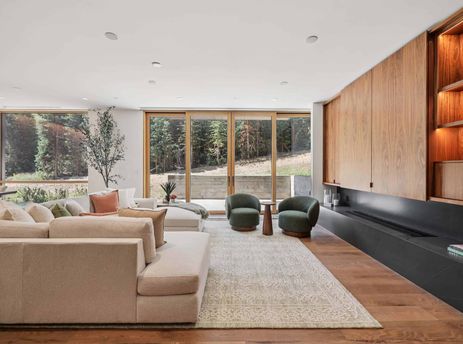 The living area at Founders Place 1403 showcases a modern design with plush seating, full-height windows offering forest views, and a stylish fireplace integrated into wooden paneling.