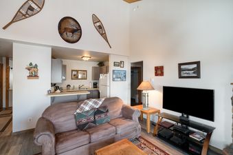 A cozy open-concept living room with a brown sofa and a kitchen area featuring modern appliances at The Pines Condominiums A304.