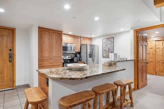 A well-lit kitchen featuring light wood cabinets, granite countertops, stainless steel appliances, and a breakfast bar with three wooden stools.