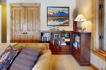 A cozy reading nook in Rendezvous D3 with a wooden bookcase, comfortable sofa, and framed mountain art.