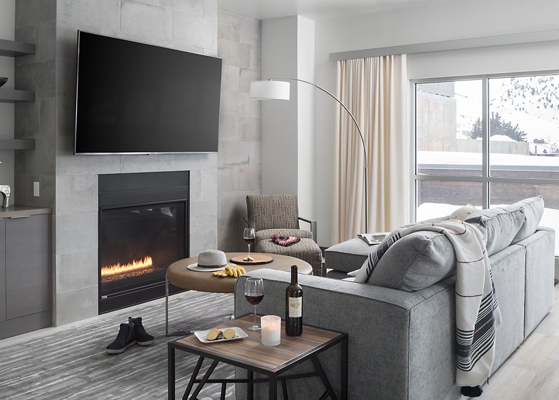 A modern living room at Penthouse on Glenwood 407 featuring a grey sectional sofa, fireplace, and flat-screen TV, with large windows overlooking a snowy landscape.