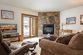 A cozy living room in Pines Condominiums Two Bedroom with a stone fireplace, comfortable seating, and a flat-screen TV, leading to a snowy patio.
