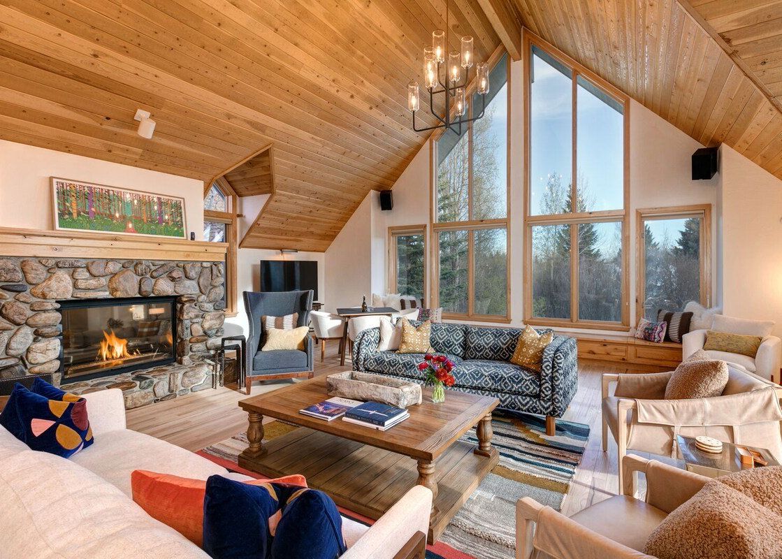 Warmly lit living room at Golden Spruce Jackson Hole featuring a large stone fireplace, vaulted wood ceilings, and tall windows looking out to the forest.