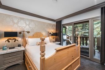 A cozy bedroom with a wooden bed and white bedding, two nightstands with lamps, and a balcony entrance with a view of lush trees.