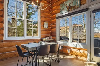A dining area with a wooden table and black chairs, surrounded by log walls and large windows offering a view of trees and buildings.