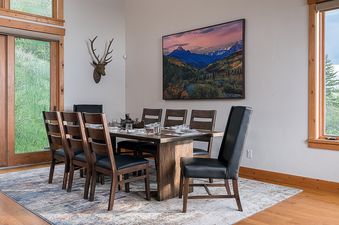 Dining area with a large wooden table and chairs, adjacent to big windows showing scenic mountain views.