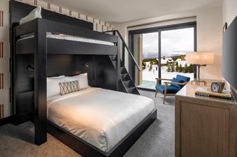 The Montage Inn Vista View Two Bedroom Residence features a bunk bed with a black wooden frame and a white duvet cover in the bedroom. There is also a large window that overlooks a snow-covered mountain view with trees and buildings in the distance.