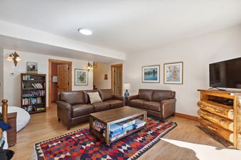 The spacious living area at Northwoods Escapes, featuring two brown leather sofas, a wooden coffee table with board games, a colorful rug, a log-style TV stand, and a bookshelf filled with books.