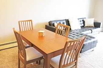 A cozy living area features a wooden dining table with four chairs and a black leather sectional sofa with throw pillows.