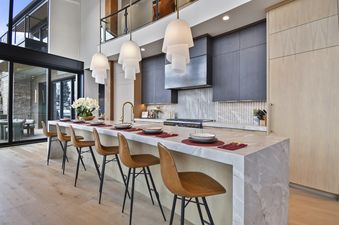 A modern kitchen with a large marble island, six brown leather bar stools, dark wood cabinetry, and unique layered pendant lights.