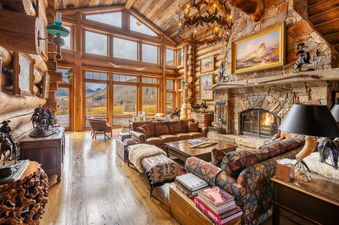 The grand living room of Elk Mountain Lodge features a stone fireplace, leather sofas, and large windows revealing an autumn mountain landscape.