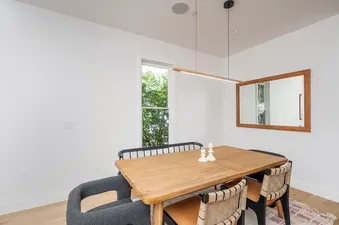 A bright dining room with a wooden table, mixed chairs, a linear pendant light, and a large mirror reflecting the room and outdoor greenery.