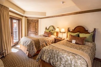 A cozy bedroom with two beds, each adorned with elegant bedding and throw pillows, complemented by a bedside table with lamps and a door leading to a balcony.