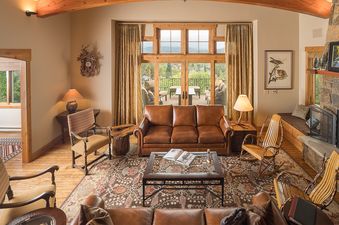 A cozy and elegantly furnished living room at the Valley View Retreat features a large leather sofa, a stone fireplace, and a glass door opening to a balcony with mountain views.