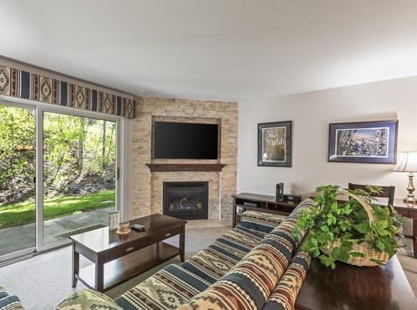 A cozy living room at Snow Flower 135 Park City featuring a comfortable sofa, a dark wooden coffee table, a stone fireplace with a mounted television, and large sliding glass doors opening to a green outdoor view.