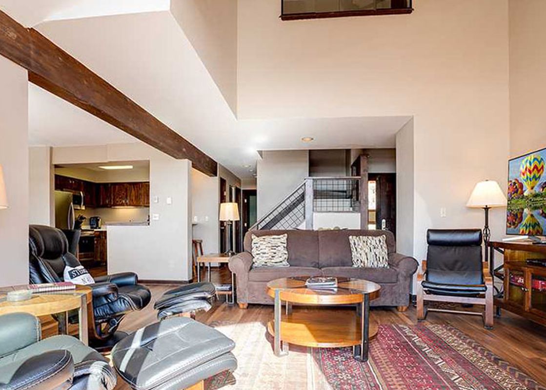 A spacious living room at Ranch at Steamboat RA106, featuring a brown sofa, leather armchairs, a large TV, and a view into the kitchen, illuminated by natural light and accented with wooden beams.