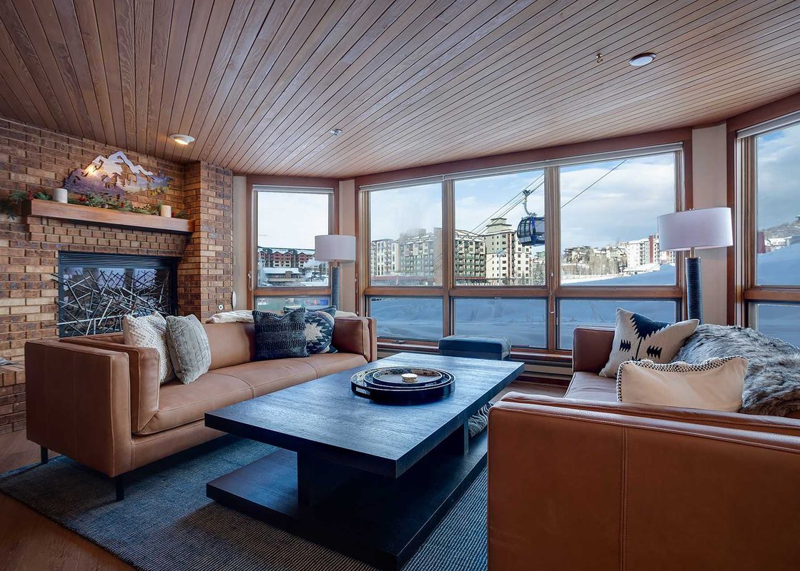 A cozy living room in Chateau Chamonix CX114 with brown leather sofas, a brick fireplace, and large windows overlooking a snowy mountain resort.