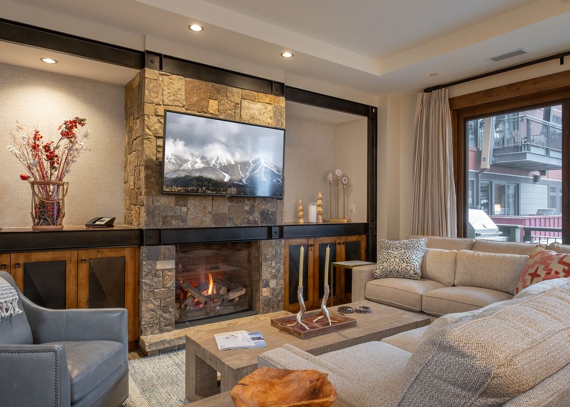 A cozy living room in Buck Mountain 2 Bedroom One Steamboat Place 307 with a stone fireplace, mounted TV, comfortable sofas, and large windows revealing a winter outdoor scene.