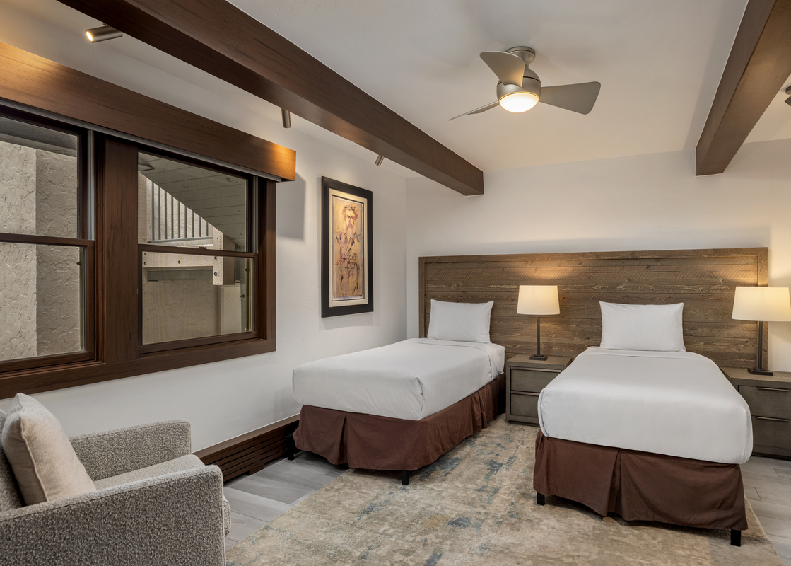 A serene twin bedroom at Top of the Village Leaf 501 with two beds, wooden headboard, modern lamps, a ceiling fan, a window, and an armchair.