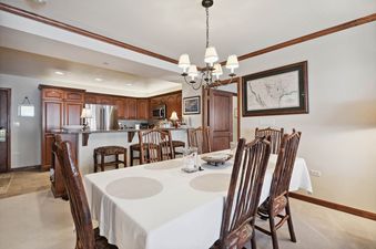 A dining room featuring a wooden dining set with a white tablecloth, a chandelier, and a view into a kitchen with wooden cabinets and stainless steel appliances.