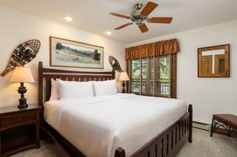 A cozy bedroom with a king-sized bed, rustic decor including snowshoes, and a window with wooden blinds offering a view of trees.
