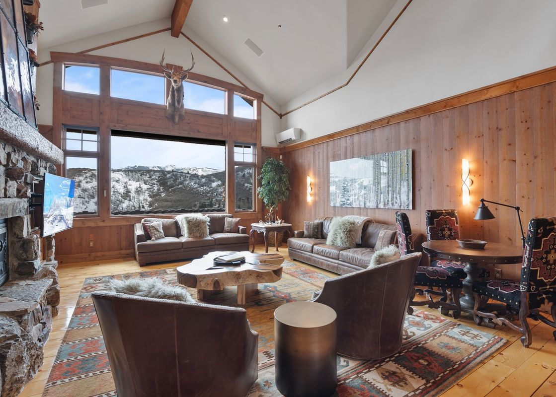 A spacious and inviting great room at the Timber Lodge at the Oaks, featuring comfortable leather seating, a stone fireplace, and large windows with views of snow-covered mountains.