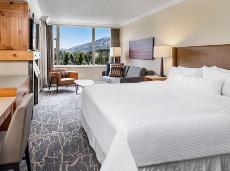 A luxurious and spacious hotel suite with a large bed, a comfortable sofa, an armchair, a desk, and a large window offering a stunning view of snowy mountains and a forest.