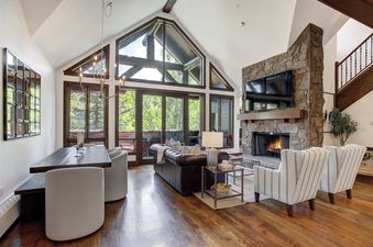 The grand living area features a stone fireplace, dark wood dining table, leather sofa, and large windows with an exterior view.