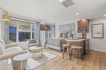 A modern studio apartment at Galleria 307 featuring a living area with a sofa and armchair, a sleeping area with a bed, and a compact kitchen island, all bathed in natural light from a large window.