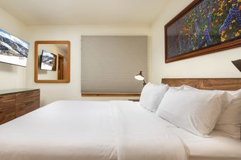 A cozy bedroom in Montaneros 212 featuring a king-sized bed with white linens, a TV, a mirror, and a vibrant panoramic artwork above the bed.