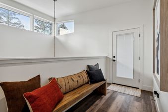 A bright and clean mudroom at Quarry A35, featuring a wooden bench with colorful pillows, large windows with tree views, and a glass-paneled exit door.