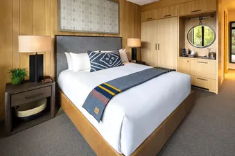 A cozy bedroom in The Sylvan Lodge Four Bedroom Penthouse with a king-size bed, nightstands, lamps, a plant, and a built-in wardrobe with a mini-bar and round mirror.