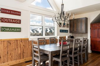 A rustic dining table with chairs is situated in the center of the room and features a rustic wood table and chairs. The table is set for a meal and has a window to the right. The background has a wooden wall and a wall with various ski-themed signs.