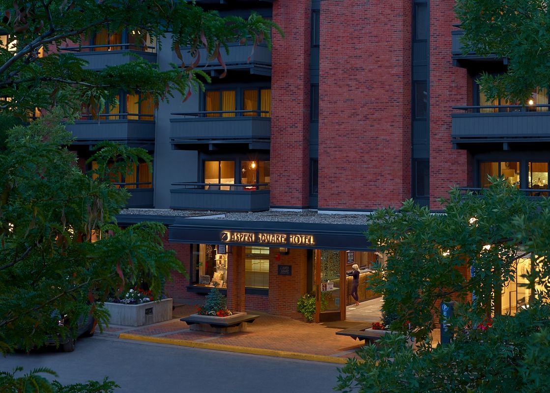 The exterior of the Aspen Square Hotel at dusk, showing the main entrance with an illuminated sign and several guest room balconies with warm lights visible through the windows.