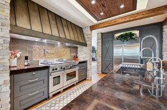The kitchen at Winter Watch Estate features a stainless steel range oven, a bronze-toned range hood, dark countertops, and light stone-clad pillars, creating a rustic yet modern culinary space.