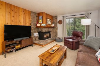 A cozy living room at Beaver Village 823 with a stone fireplace, large television, and comfortable seating, featuring a view of evergreen trees from the balcony.